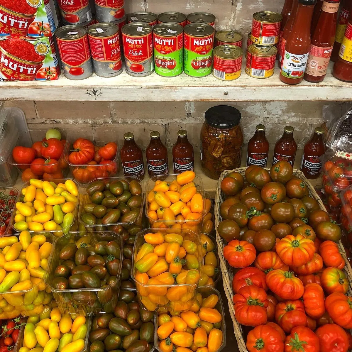 מדף בחנות עם קופסאות עגבניות שרי צבעוניות, עגבניות ותיקות, ומעליהן שימורי עגבניות